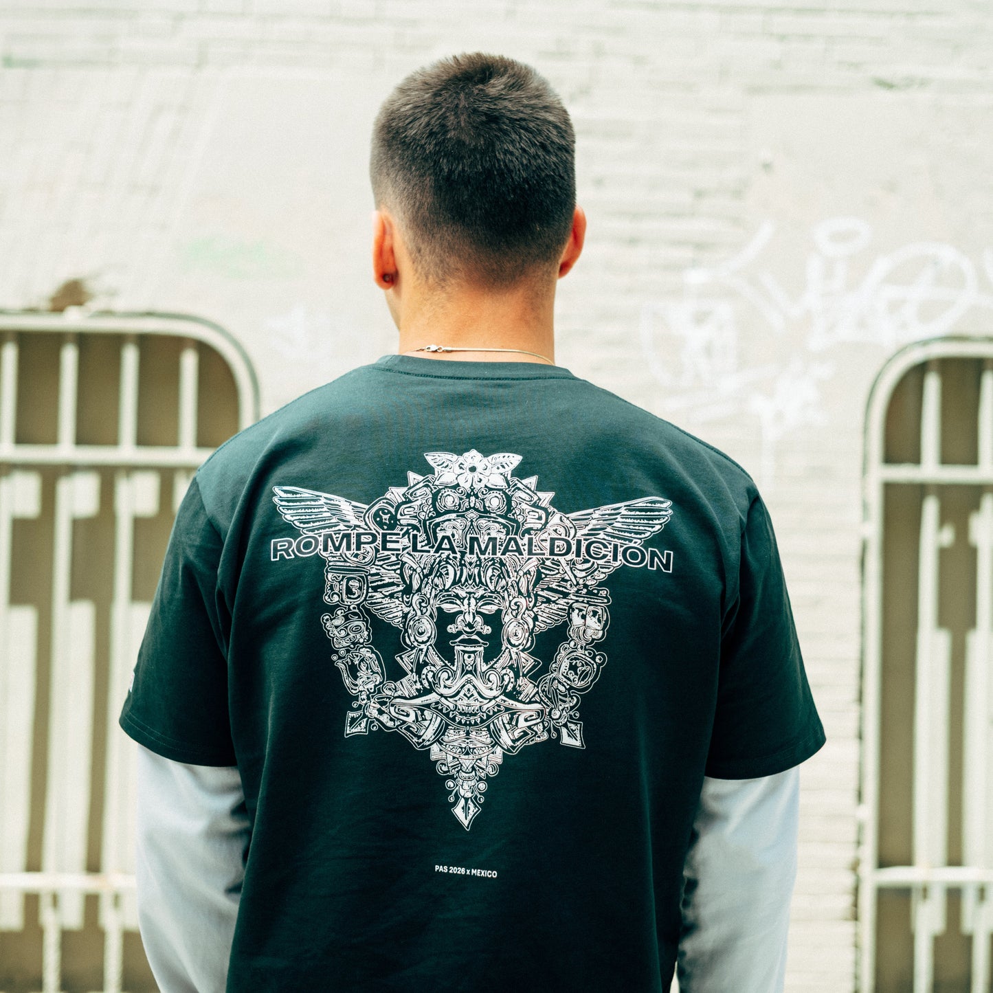 PAS x Mexico Rompe La Maldición El Tri dark green tee – Aztec back graphic with Rompe La Maldición text, worn against urban wall
