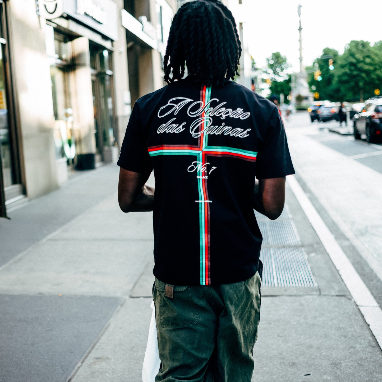 Black Portugal fútbol tee shown from the back on a model walking a NYC street, with "A Seleção das Quinas" script above a red-and-green Portuguese cross and "No. 7" graphic.