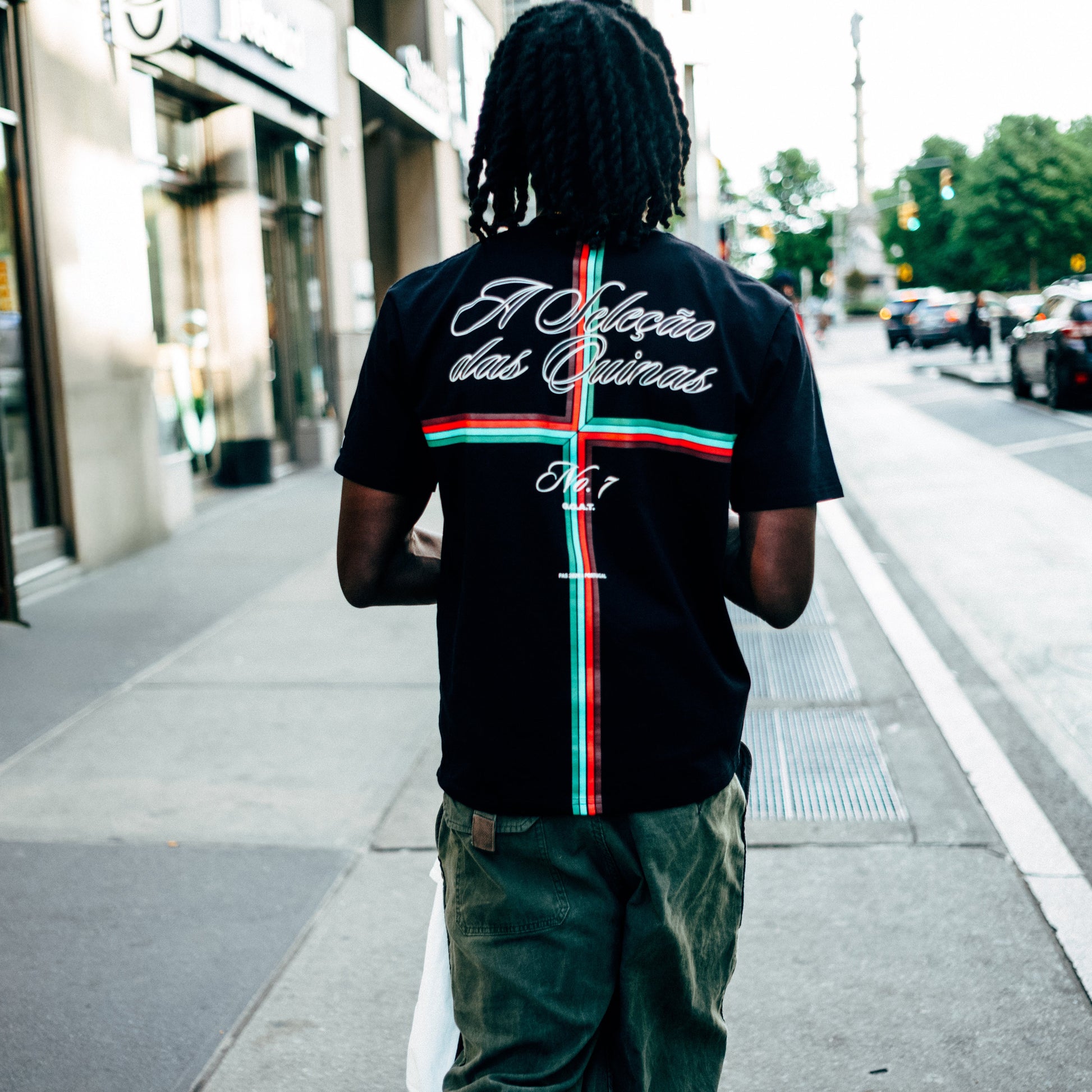 Black Portugal fútbol tee shown from the back on a model walking a NYC street, with "A Seleção das Quinas" script above a red-and-green Portuguese cross and "No. 7" graphic.